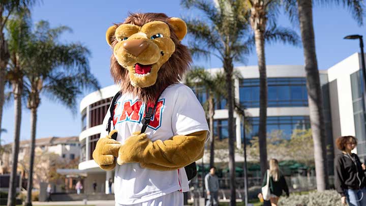 LMU mascot Iggy the Lion wearing a backpack with the William H. Hannon Library in the background