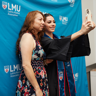 Two women taking a selfie agains the Latino Alumni Association backdrop
